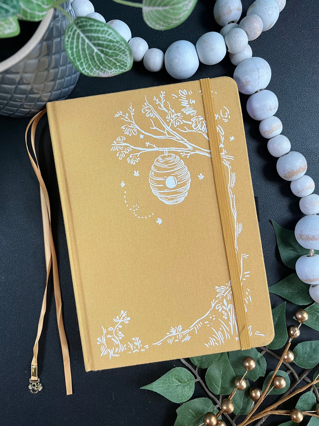 A5 Notebook from the Summer 2025 Subscription Box: Marigold color with white debossing images of tree, bee hive, bees